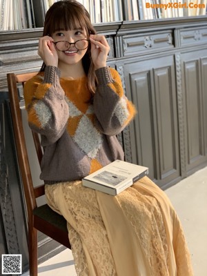 A woman sitting on a chair holding a book and wearing glasses.